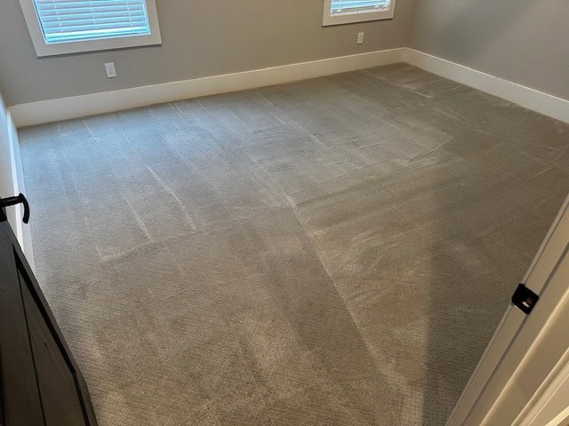 An empty room in TX with beige carpet—featuring visible vacuum lines from recent carpet cleaning Houston County—light gray walls, white trim, and two windows with closed blinds. No furniture is present.