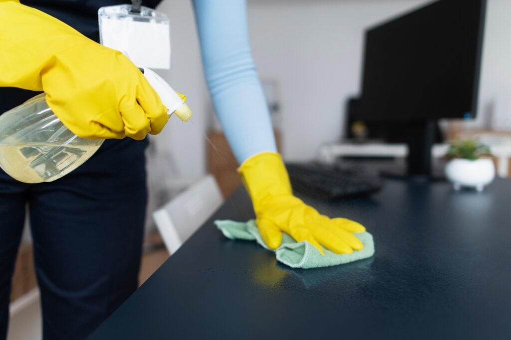 A person in yellow rubber gloves cleans a dark desk with a green cloth and sprays cleaner, with a computer monitor and keyboard visible—just another spotless scene like those from top carpet cleaning Houston County, TX services.