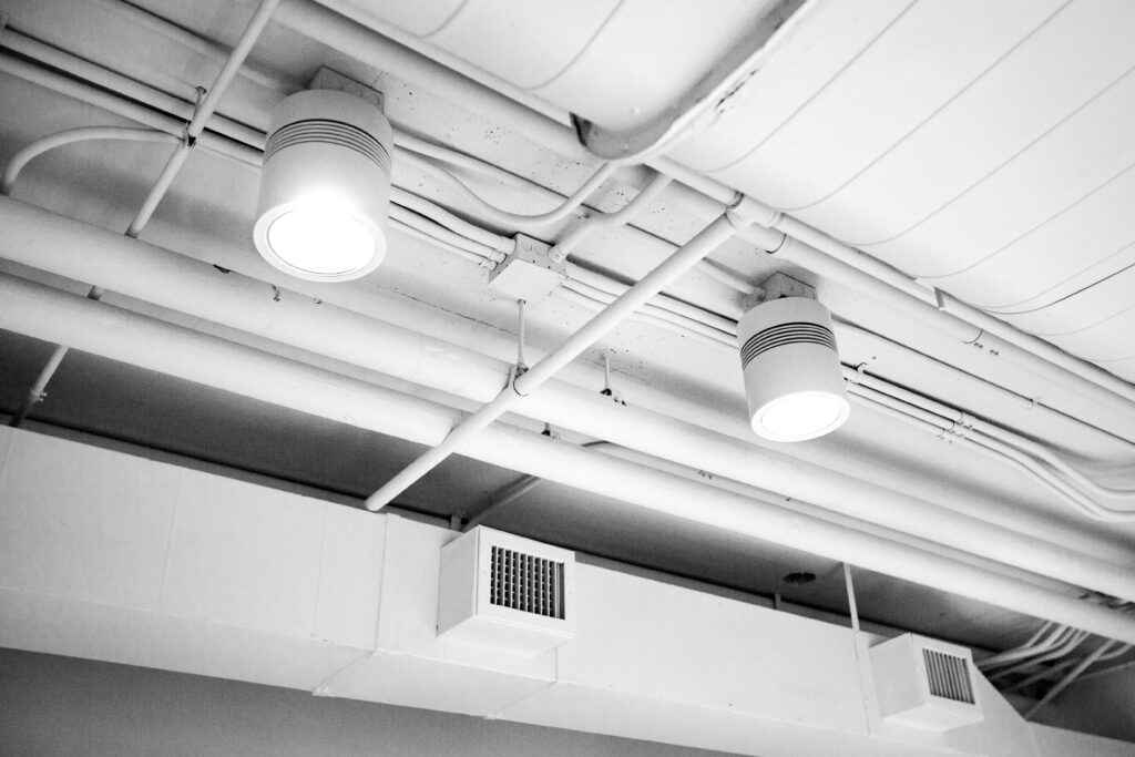 Two round ceiling lights glow among exposed white pipes and ventilation ducts on an industrial-style ceiling in TX. The photo, in black and white, echoes the clarity you expect from professional carpet cleaning Houston County services.