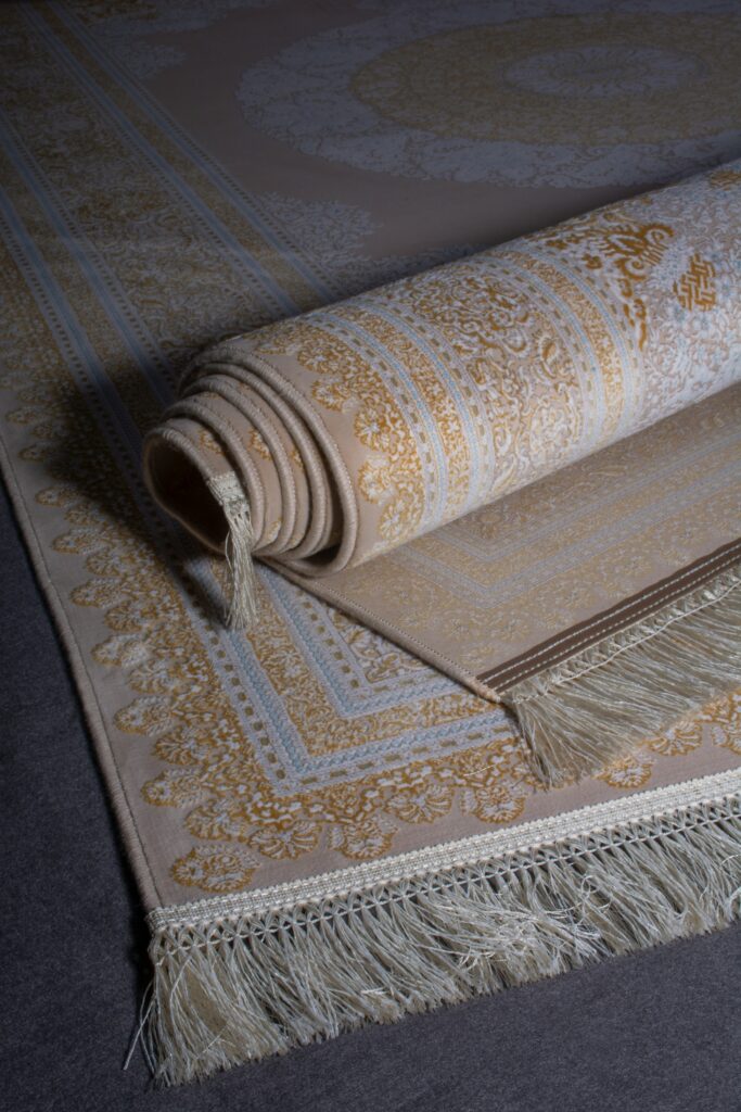 A rolled-up ornate beige and golden patterned rug with intricate designs and fringed edges lies partially unrolled on a dark surface, ready for expert carpet cleaning Houston County, TX.