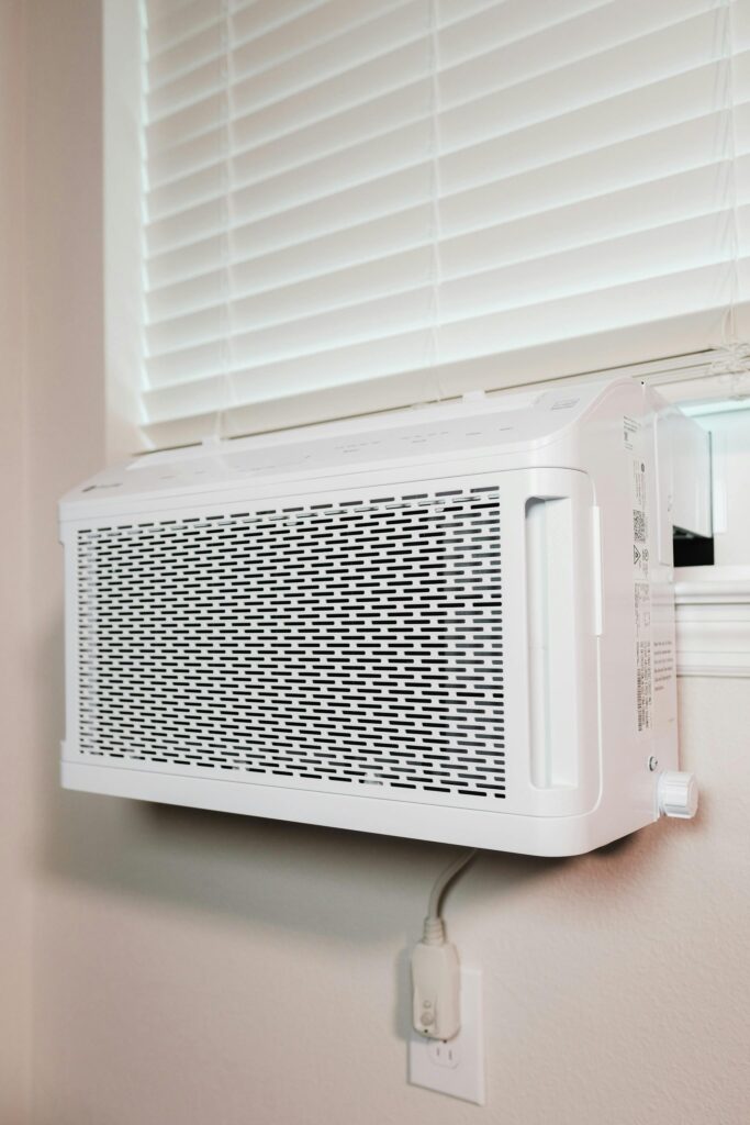 A white window air conditioner unit is installed in a window with closed blinds, above an electrical outlet. For homes in need of carpet cleaning, Houston County, TX residents can enjoy cool air and fresh floors all season long.