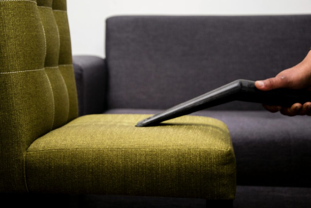 A person uses a vacuum cleaner with a nozzle attachment to clean the seat of an olive-green upholstered chair, showcasing attention to detail similar to professional carpet cleaning in Houston County, TX; a gray sofa sits in the background.