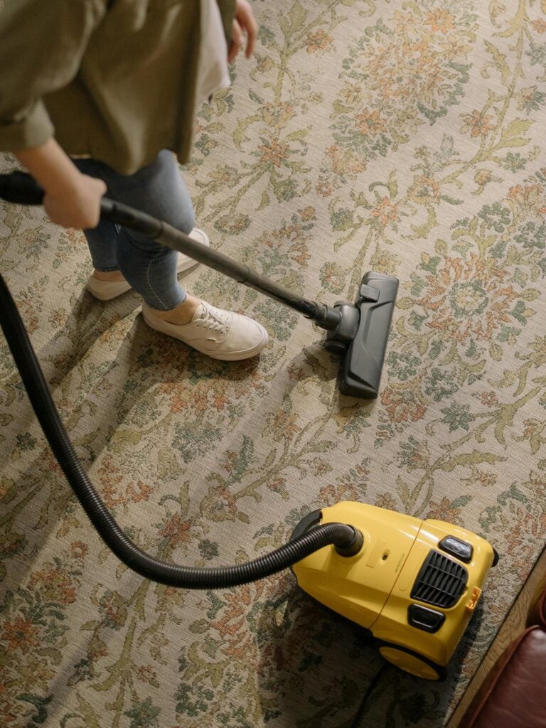 Person vacuuming a patterned carpet with a yellow canister vacuum cleaner, wearing jeans and white sneakers—ideal for efficient carpet cleaning in Houston County, TX. View from above.