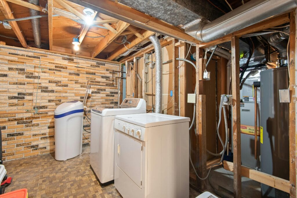 Unfinished basement laundry area in TX with exposed wood beams, brick wall, washing machine, dryer, water softener, water heater, and furnace—perfect for storage or even carpet cleaning Houston County projects.