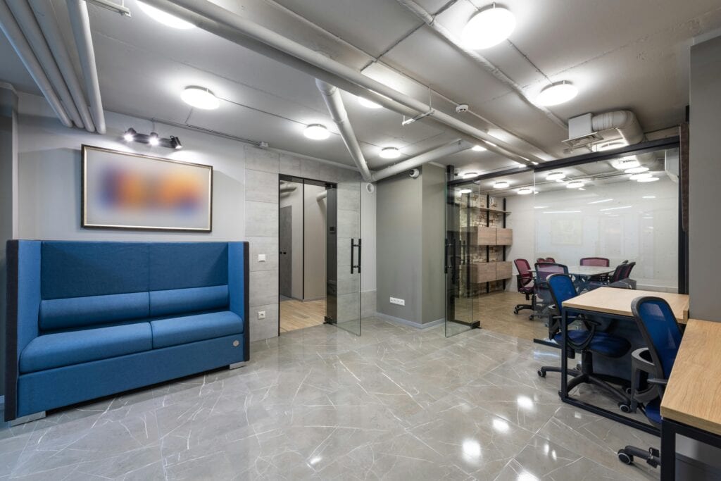 Modern office space with gray walls and ceiling, blue cushioned bench on the left, wooden desks with chairs on the right, glass-walled meeting room in the background, abstract art on the wall, and spotless floors thanks to carpet cleaning Houston County, TX.
