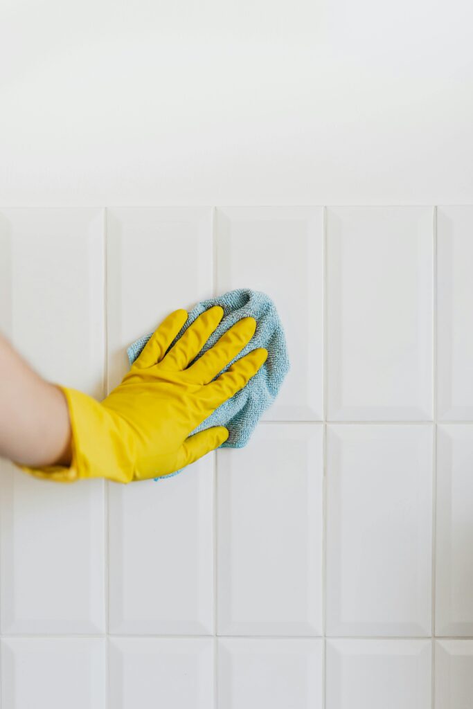 A person wearing a yellow rubber glove cleans white tiled walls with a blue cloth, showcasing the same attention to detail you'd expect from professional carpet cleaning in Houston County, TX.