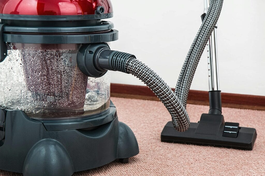 A close-up of a vacuum cleaner with a transparent water tank, flexible hose, and carpet attachment on a pink carpet near a white wall—perfect for carpet cleaning in Houston County, TX.